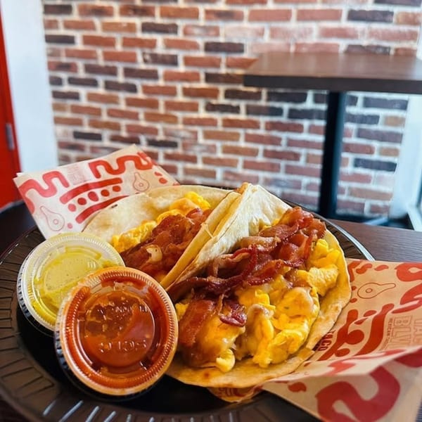 Two breakfast tacos with scrambled eggs and bacon, with two sauce cups, on a black plate.