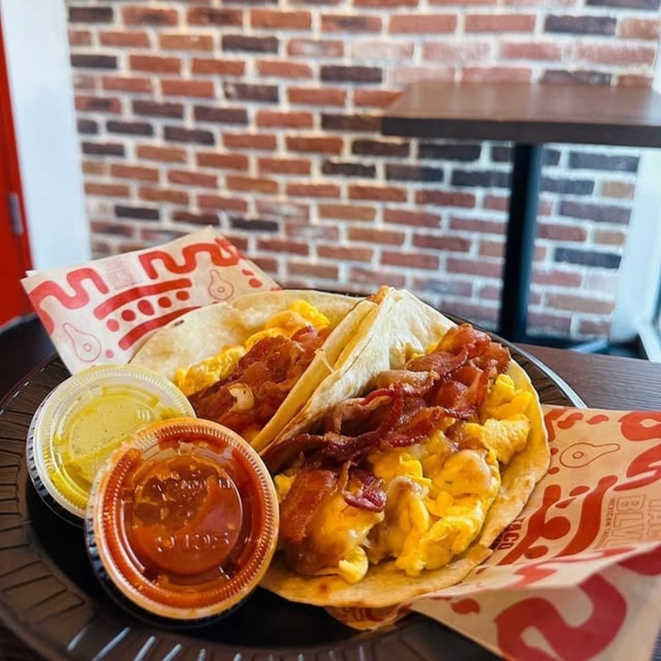 Two breakfast tacos with scrambled eggs and bacon, with two sauce cups, on a black plate.