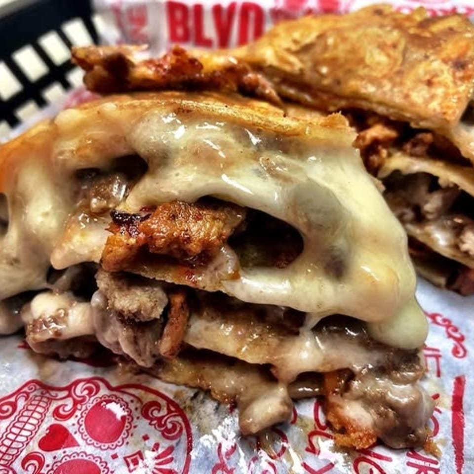 Close-up of a cheesy Taco Blvd quesadilla with meat filling, served in a basket with branded paper.