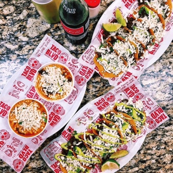 Three plates of Taco Blvd tacos and two bowls of soup, with a Coca-Cola bottle.