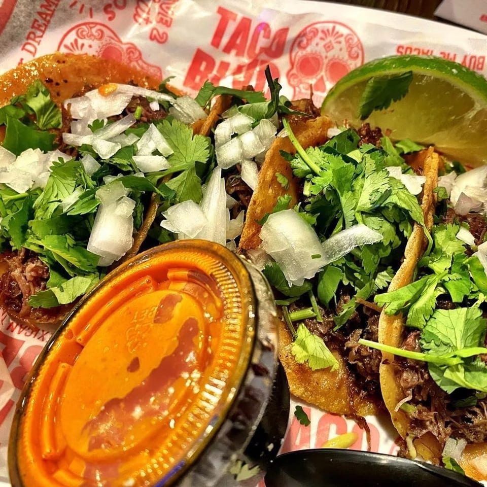 Close-up of three Taco Blvd tacos with meat, cilantro, onions, lime, and a side of sauce.