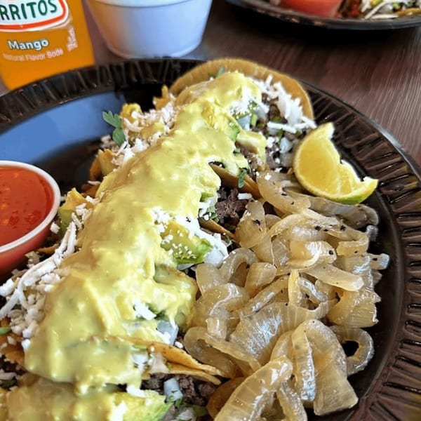 Three tacos topped with yellow sauce, cheese, and avocado, with a side of onions and lime.