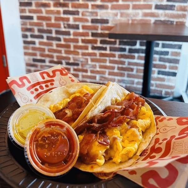 Two breakfast tacos with scrambled eggs and bacon, served with salsa and sauce cups.