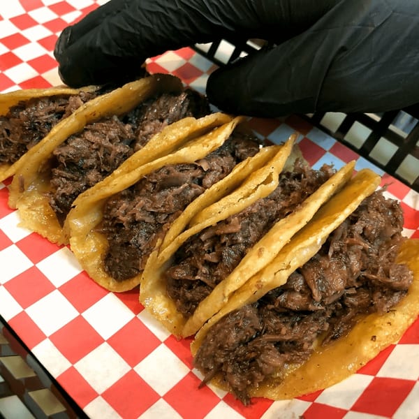 A gloved hand places a Taco Blvd taco with shredded meat into a basket lined with red and white checkered paper.